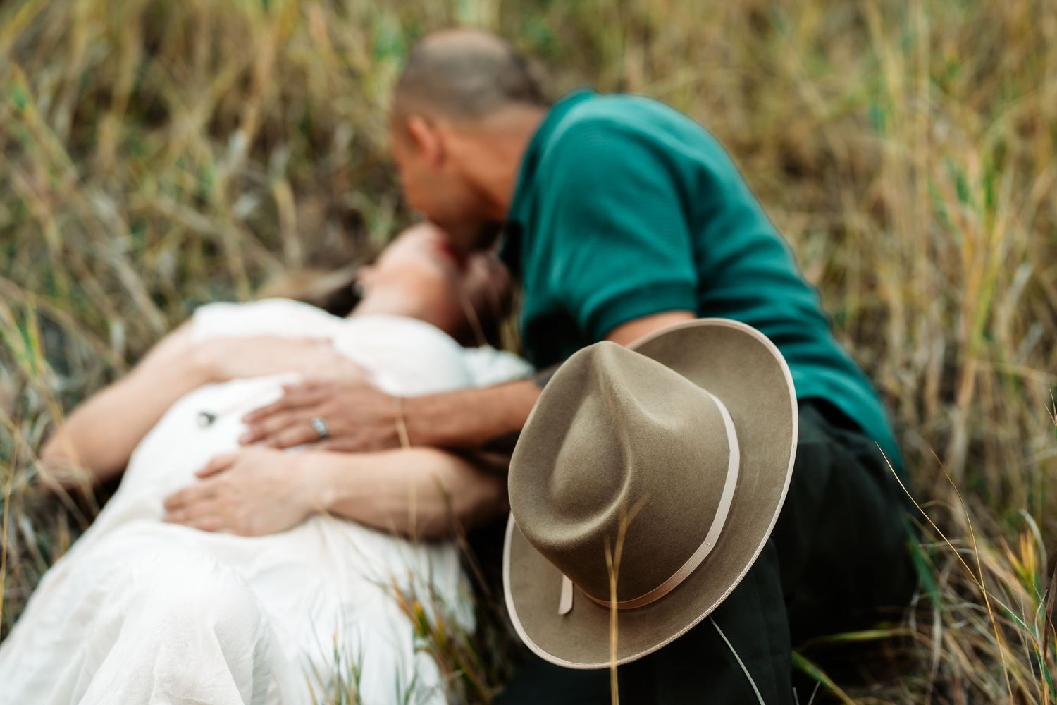 A person in a green shirt lies in tall grass while petting a white and brown spotted dog outdoors.