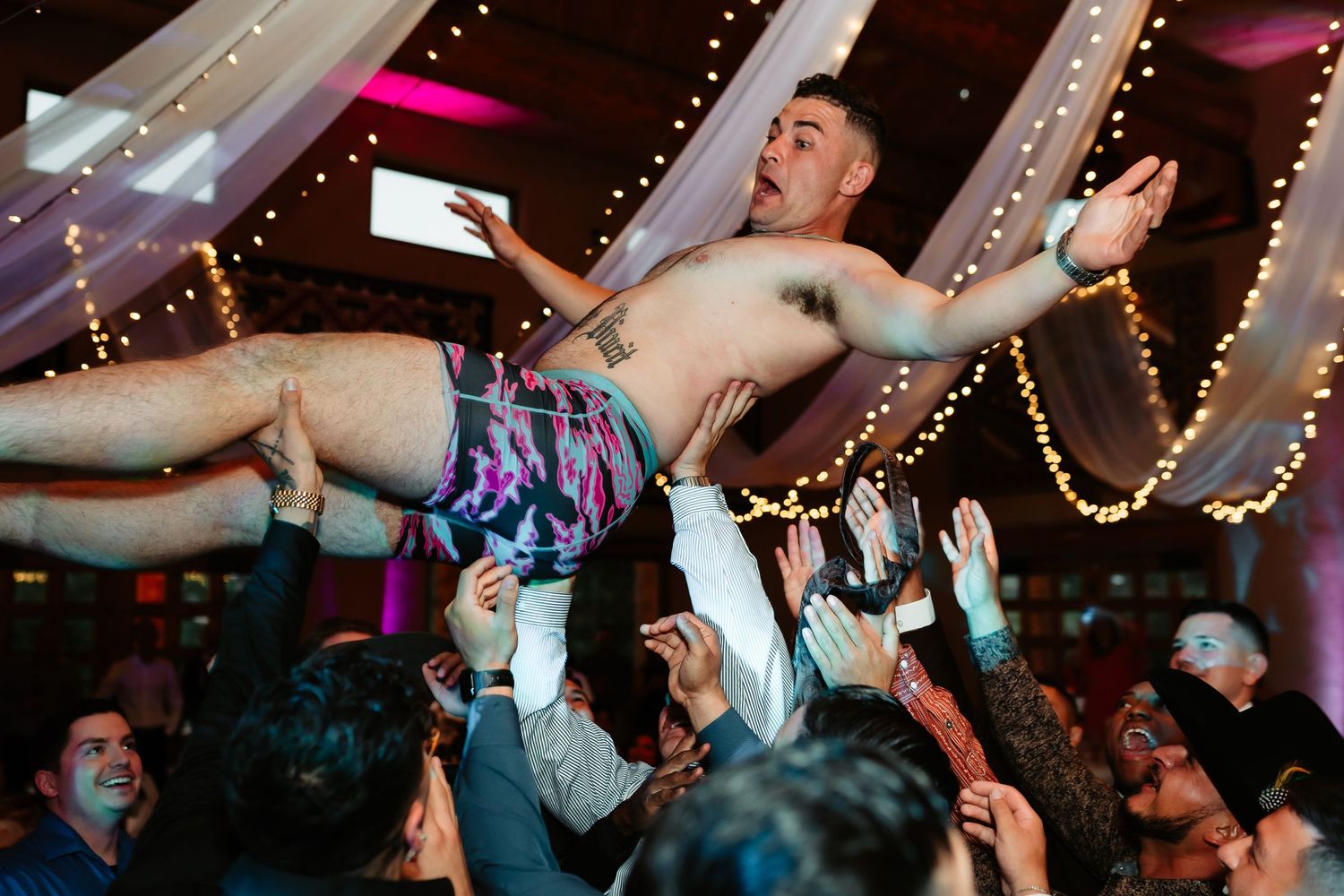 A crowd of people dancing with raised arms at a lively party with string lights overhead in the venue.