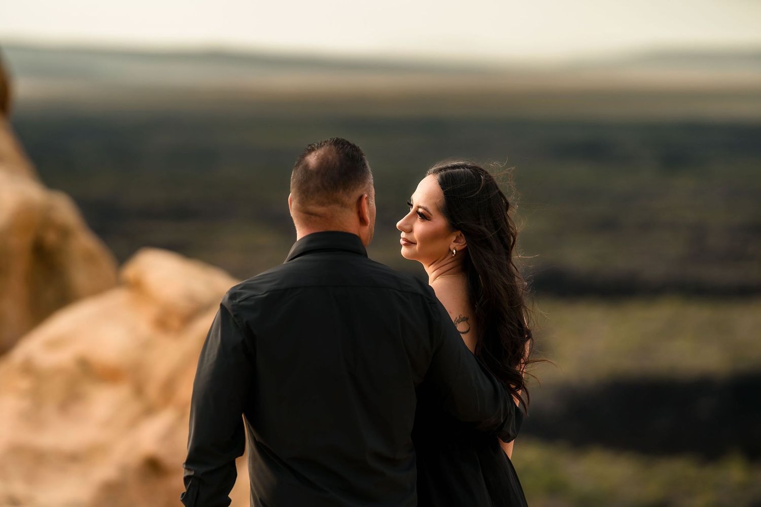 Romantic couple dressed in black sharing an intimate moment on a cliff with a vast landscape in the background.