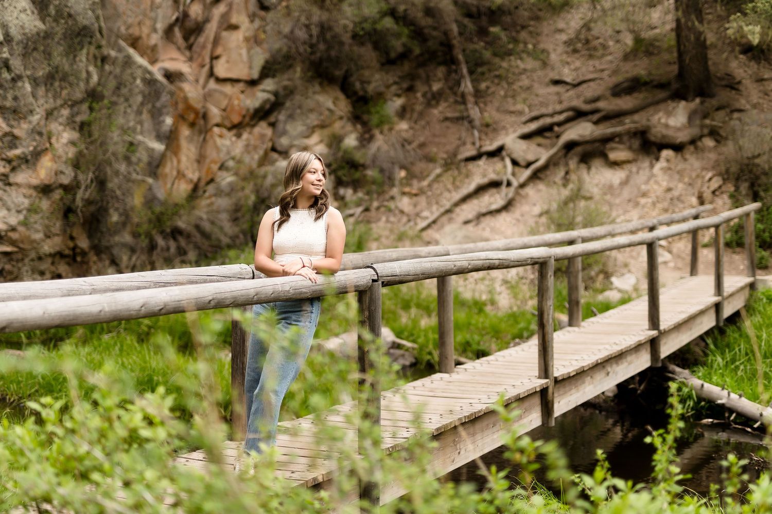 Young woman in casual outfit leans on wooden bridge railing surrounded by lush green nature.