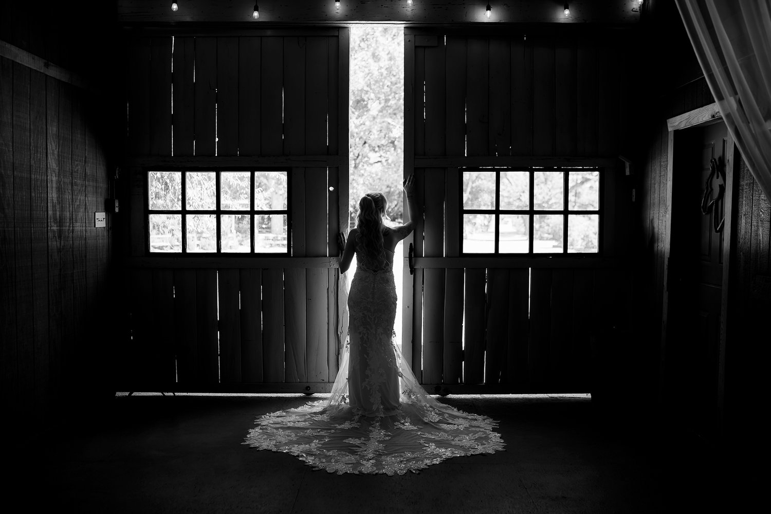 Bride in lace wedding gown stands silhouetted before barn doors in elegant black and white bridal portrait.