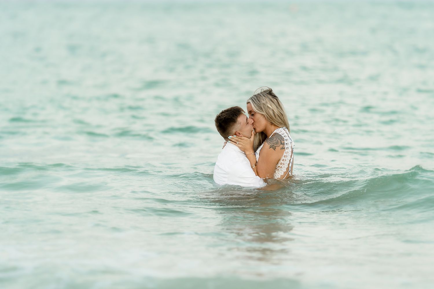 Couple sharing a romantic kiss while standing in the ocean waves during a beach engagement photo session.