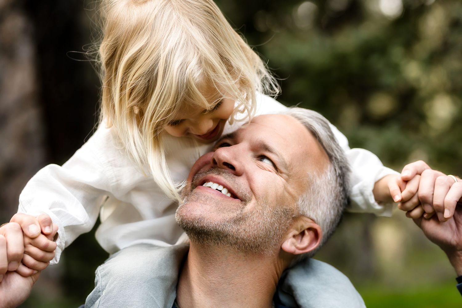 A happy father smiles while his young blonde daughter sits on his shoulders and kisses his cheek outdoors.