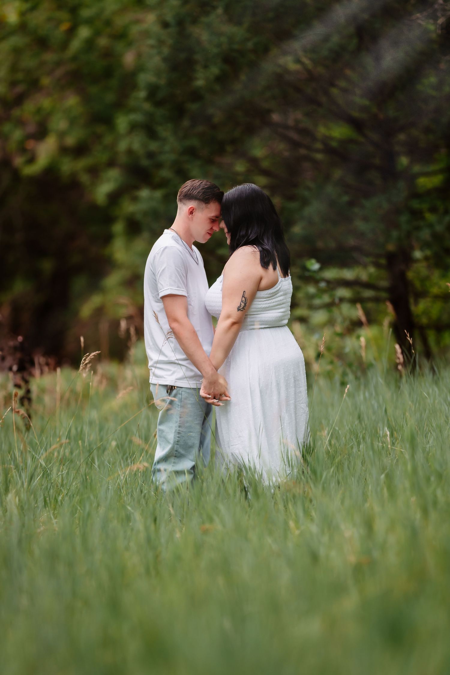 Couple shares intimate moments while standing in a tall grass meadow with trees in the background at dusk.