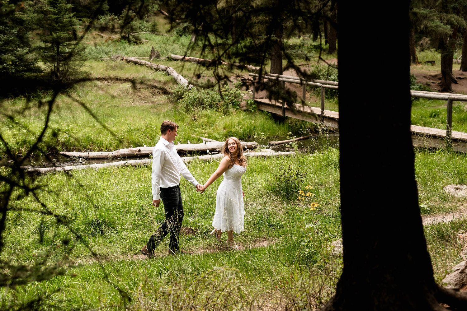 A couple in wedding attire holds hands in a lush green forest meadow with fallen logs in the background.