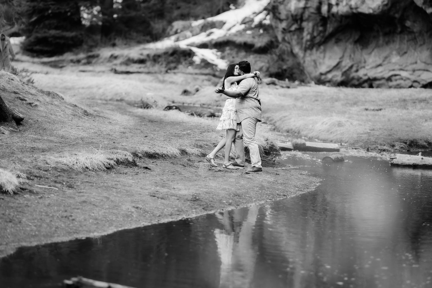 Black and white image of couple embracing by reflective water's edge in natural setting.
