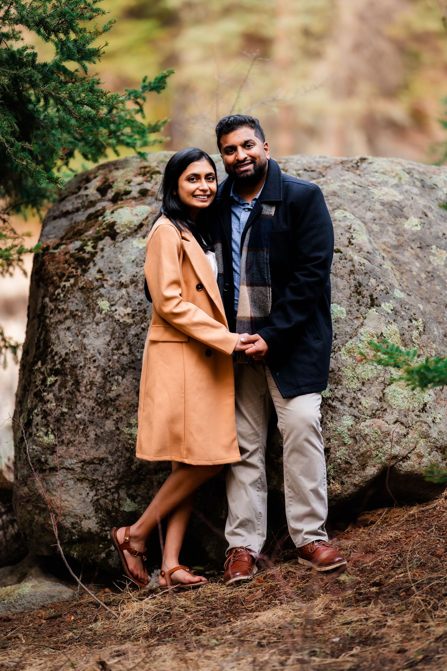 A couple poses together against a large boulder in an outdoor setting with natural scenery.