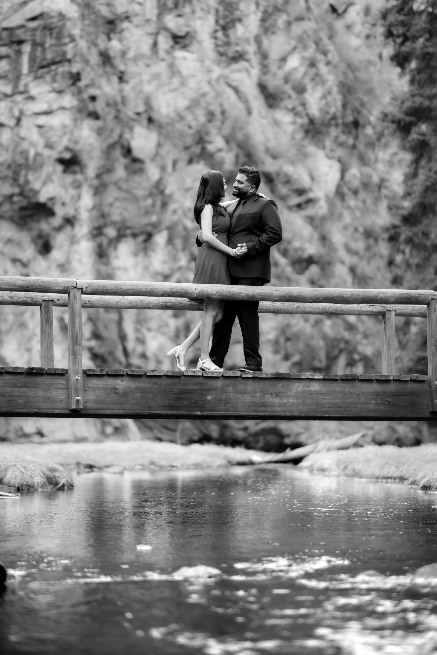 Couple stands together on bridge over stream with rocky cliff backdrop in black and white.
