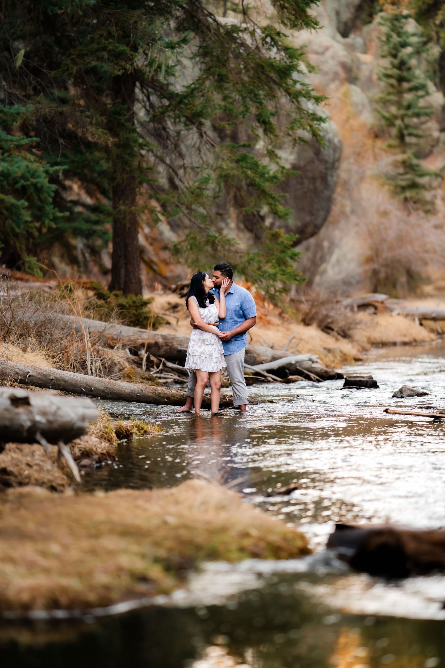 A couple embraces by a mountain stream with rocky cliffs and evergreen trees in the background.