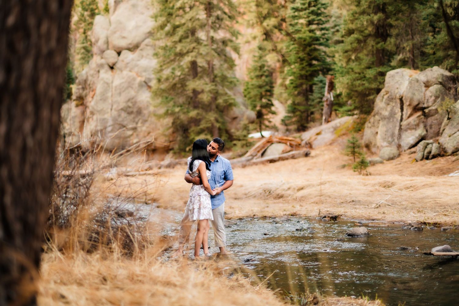 A couple shares an intimate moment by a stream with rocky cliffs and pine trees in the background.