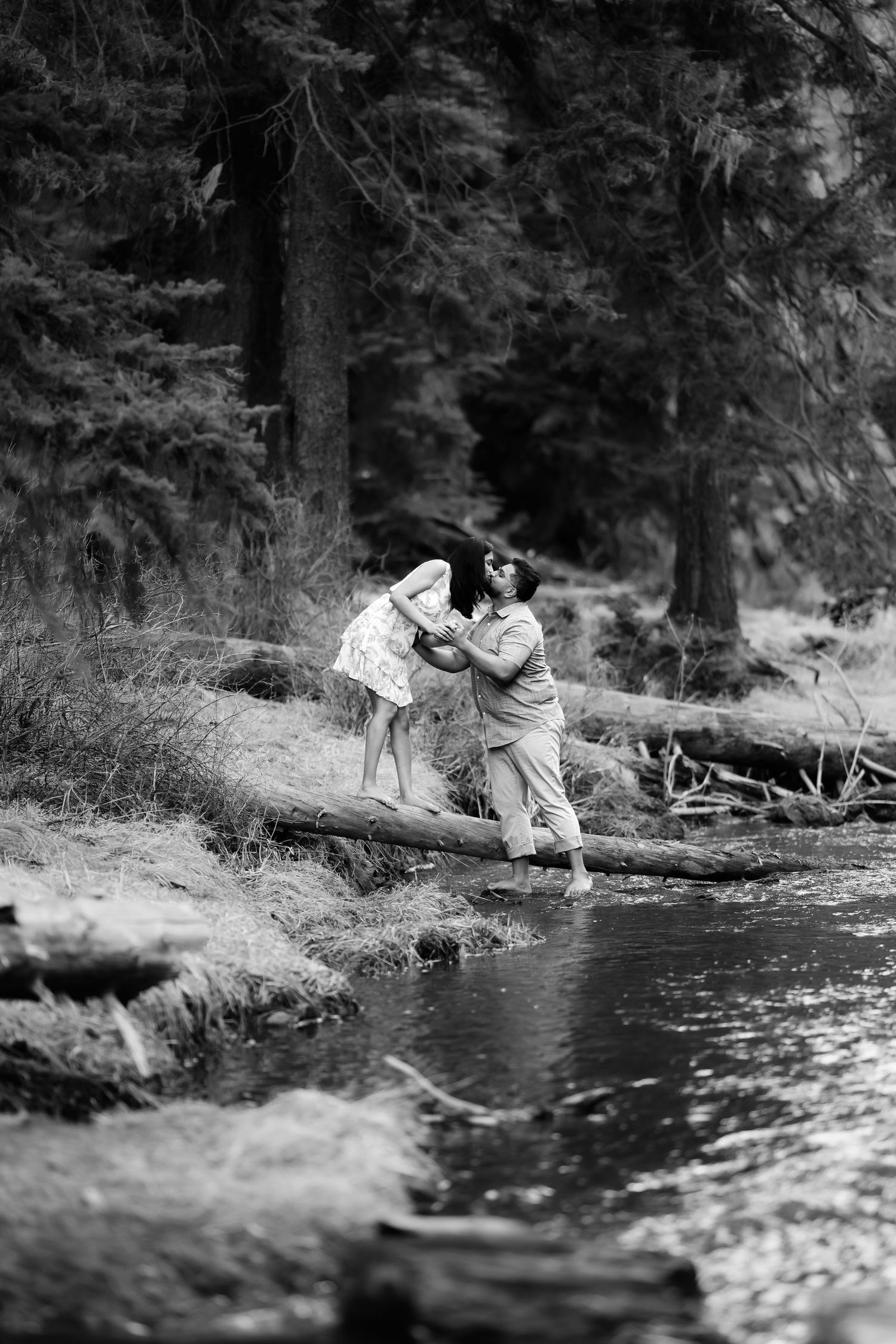 A couple balances playfully on a fallen log over a rushing stream in a forest setting.