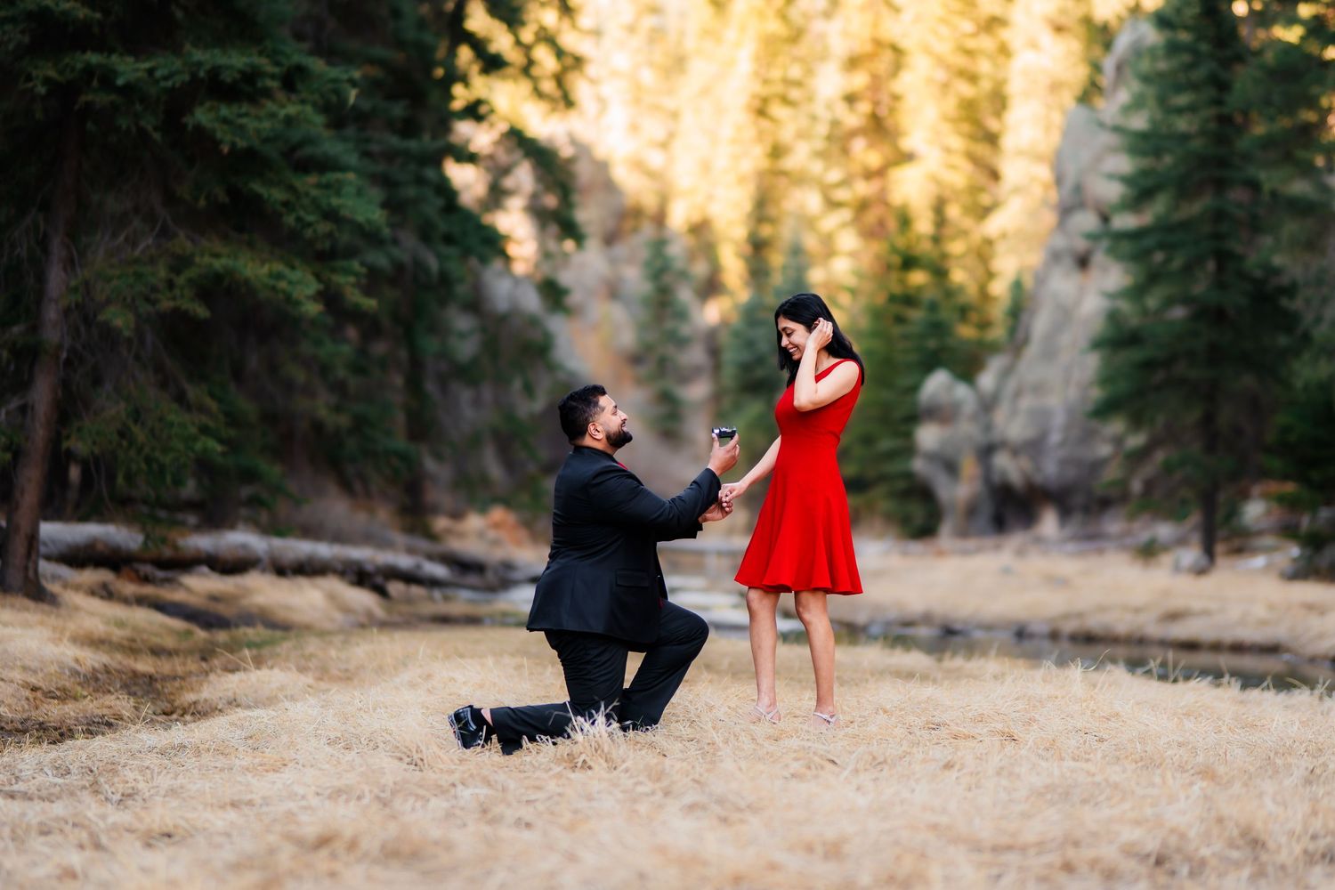 A couple sharing a romantic proposal moment by a lake with pine trees and rocky cliffs at sunset.