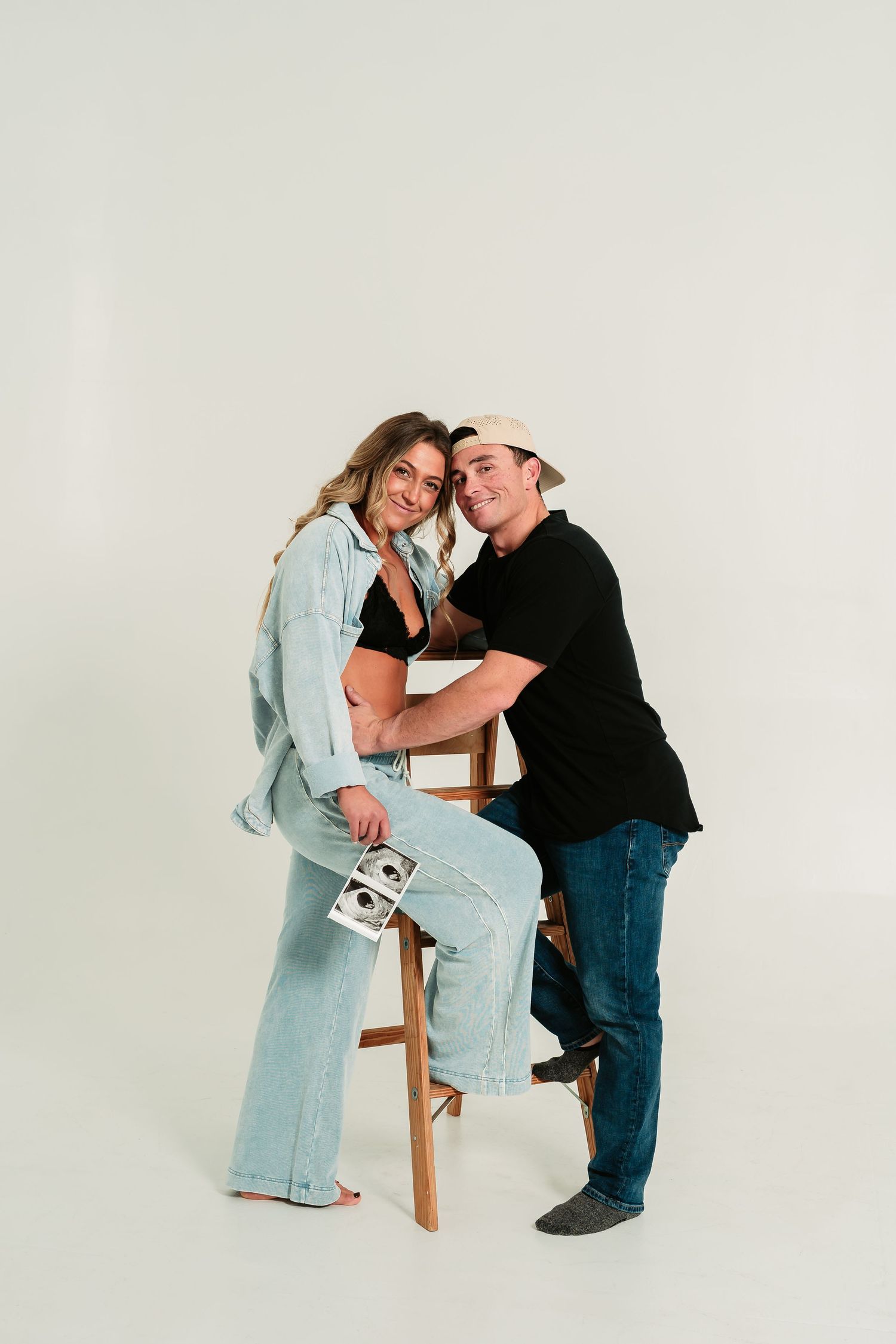 A couple in casual attire shares intimate moments in a minimalist white studio setting.