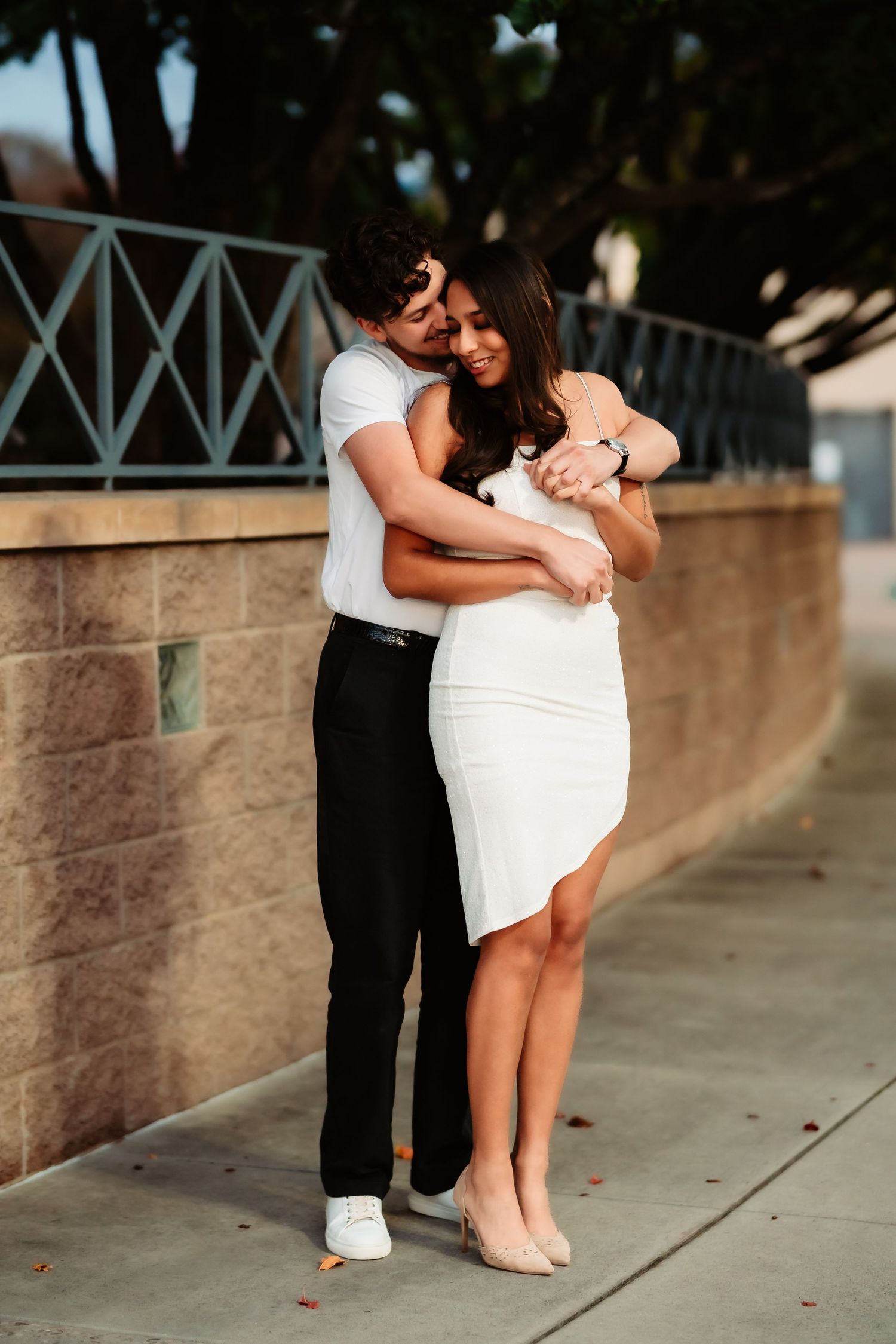 A couple shares an intimate moment while standing against a stone wall with white railings.