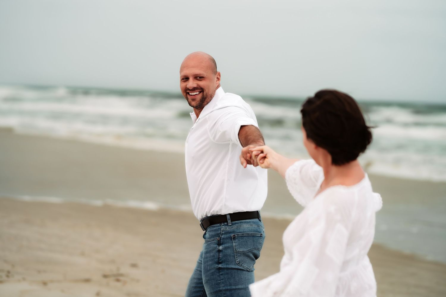 Two people in white shirts share a joyful moment together on a cloudy beach.