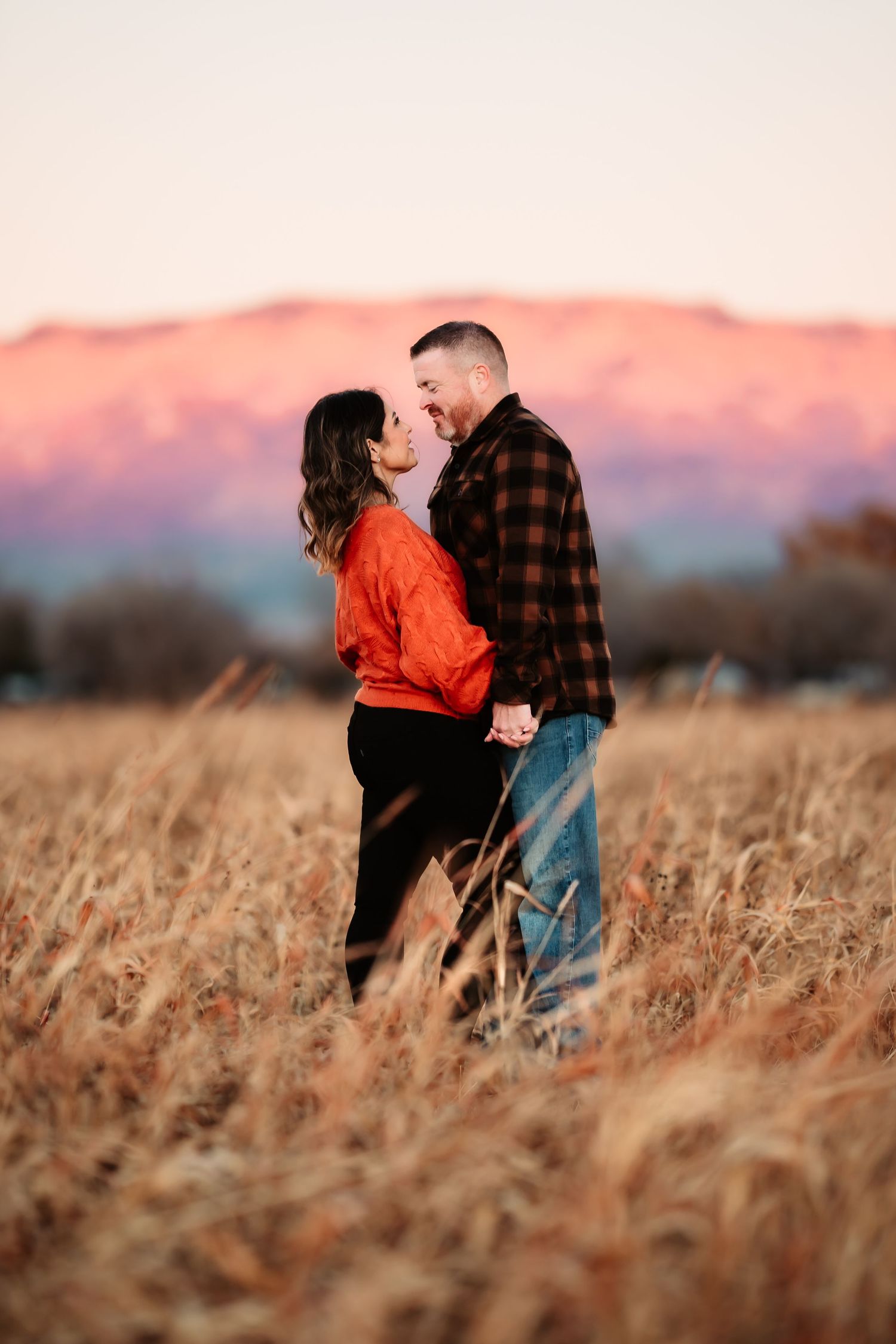 A couple embraces in a golden wheat field during sunset with pink and purple skies in the background.