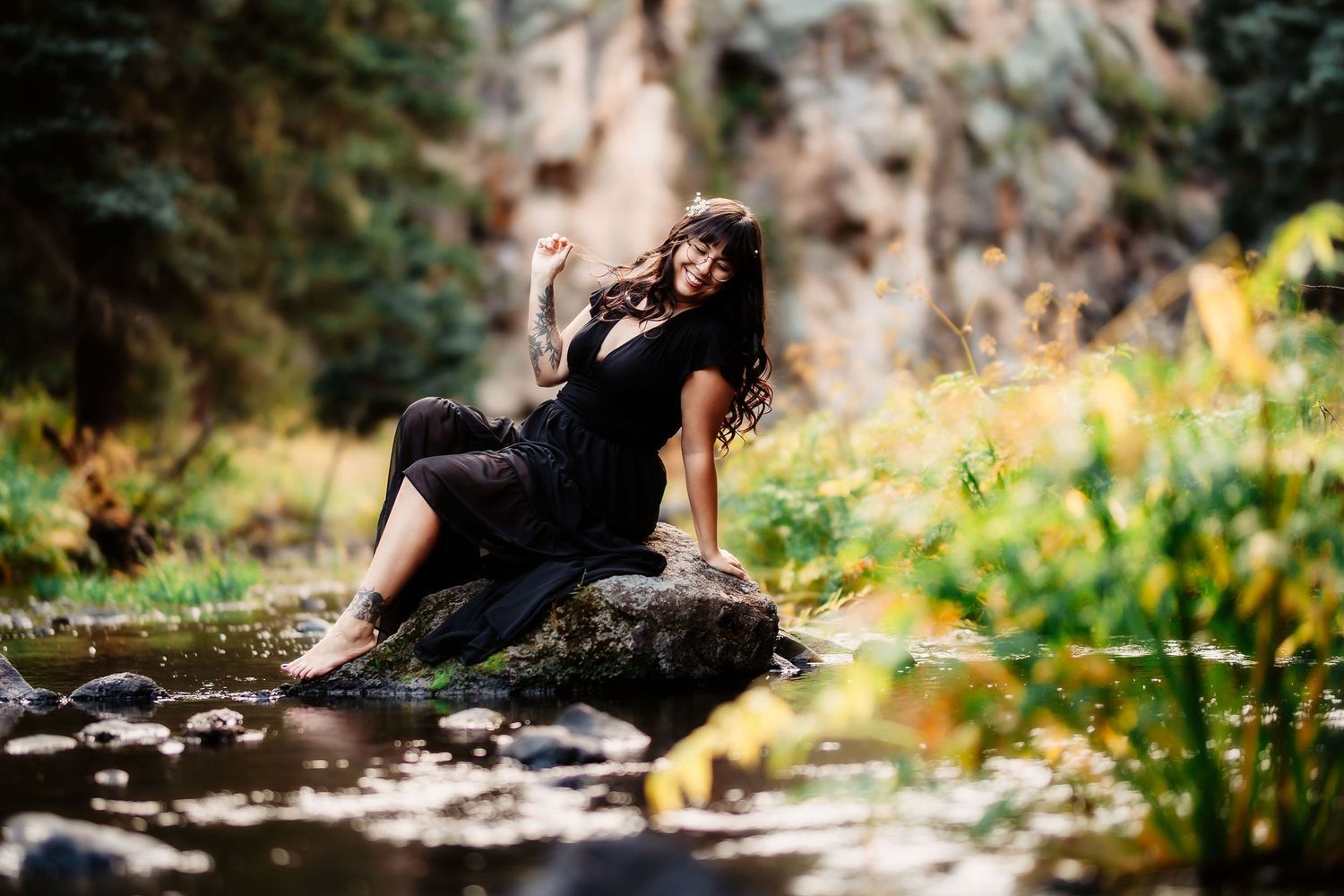 Woman in black dress sitting gracefully on rock beside tranquil forest stream in nature.