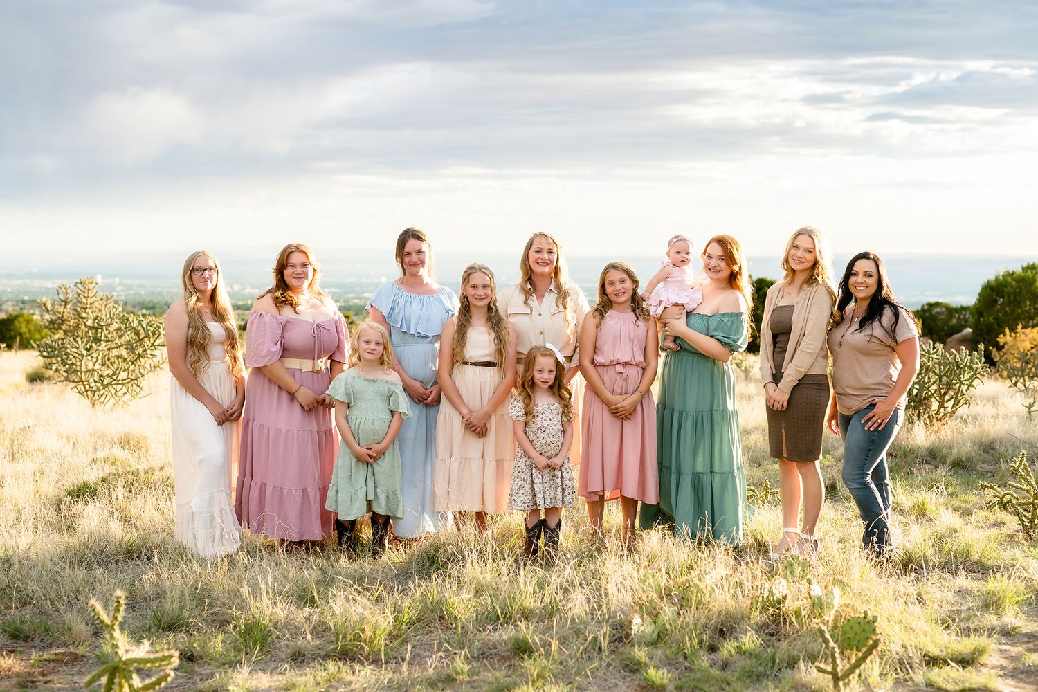 Group of women and girls in pastel dresses posing outdoors in a field with a scenic sky backdrop.