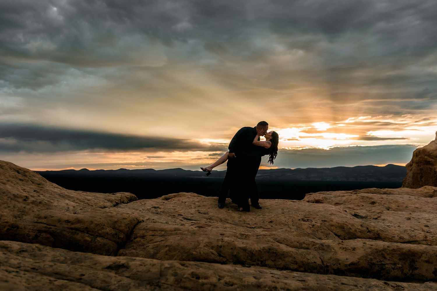 A couple shares a passionate dip kiss on desert rocks during a stunning golden sunset with dramatic clouds.