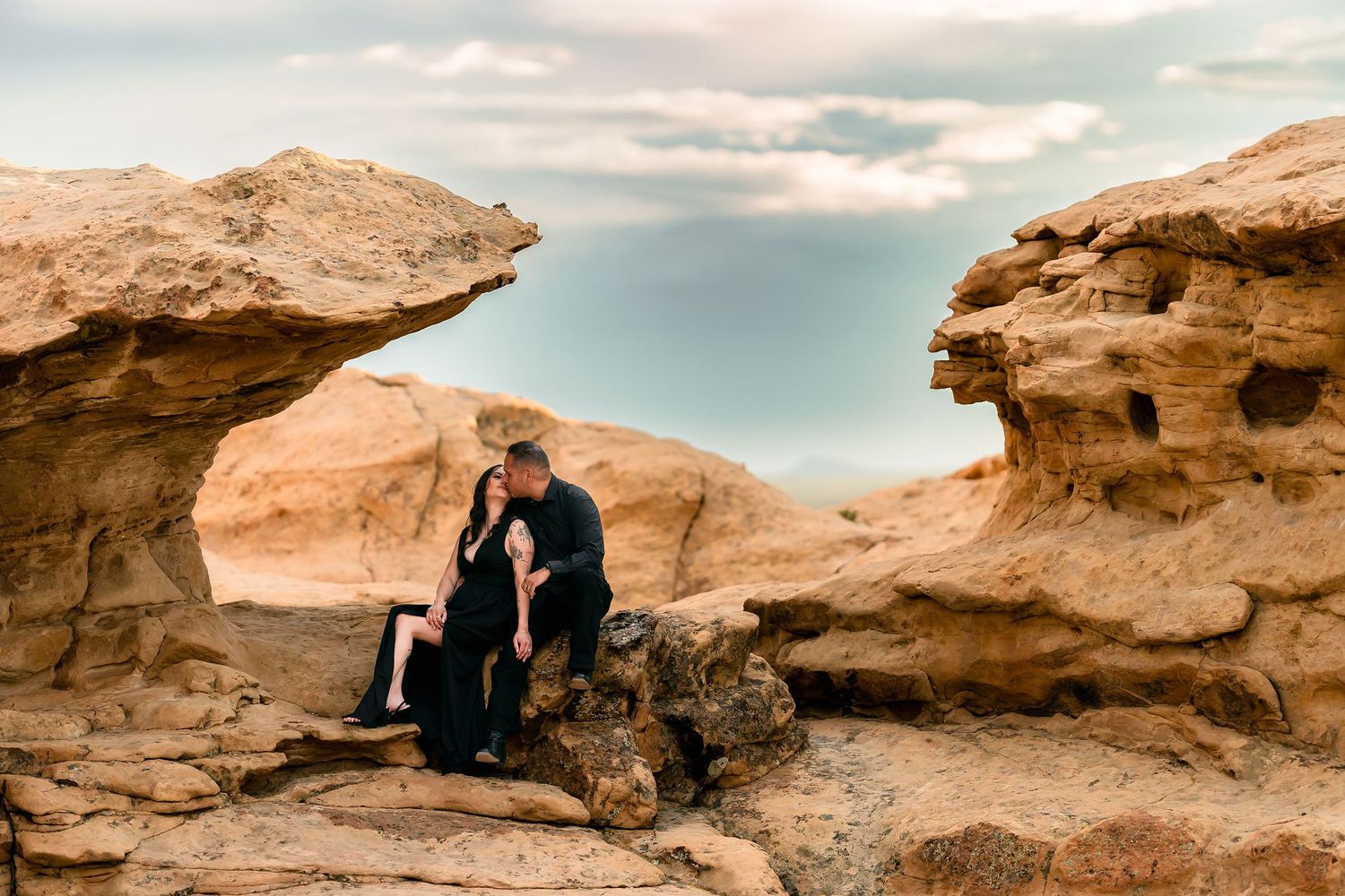 Couple in black sitting intimately between dramatic sandstone rock formations under cloudy sky.