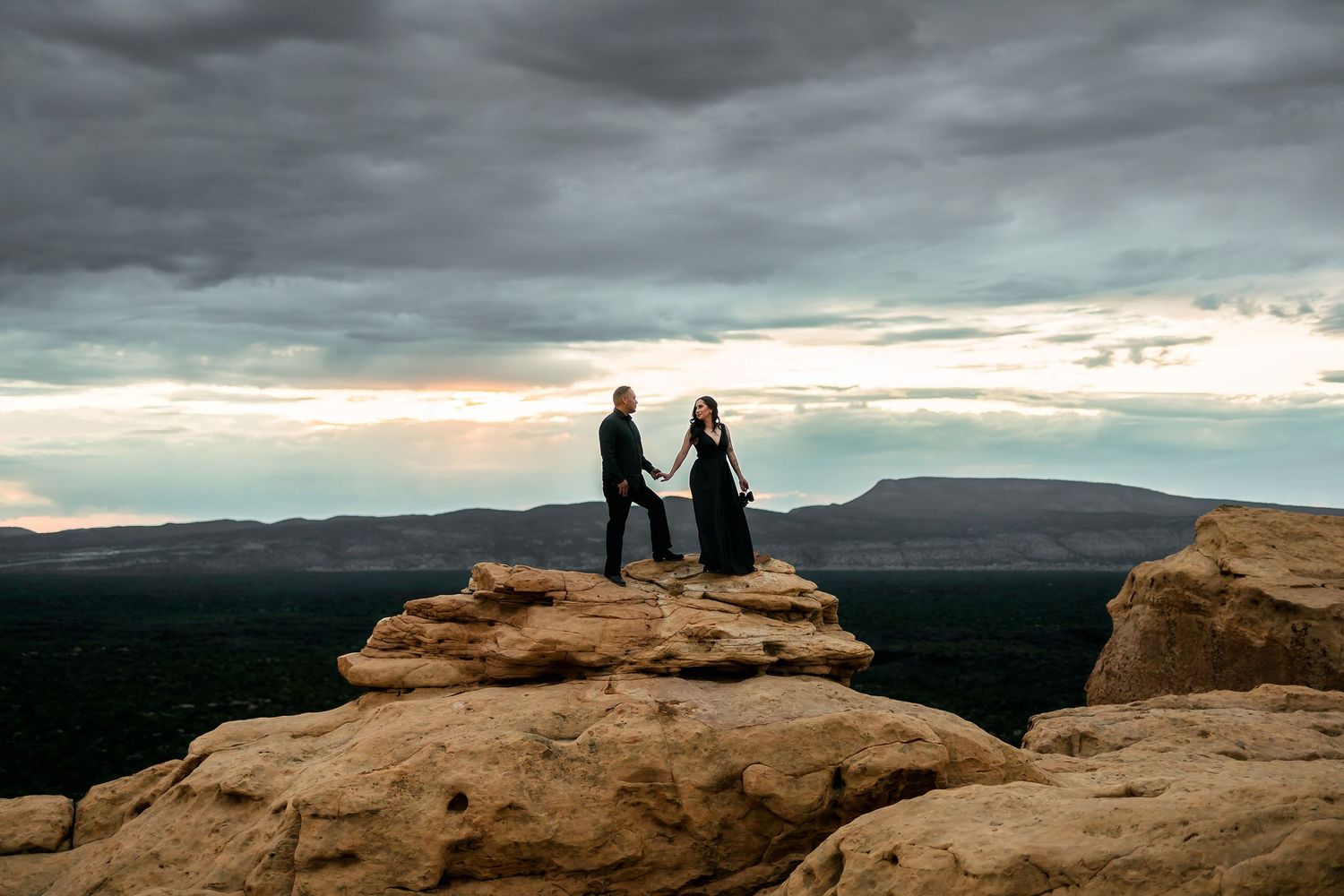 A couple holds hands atop a rocky cliff under dramatic stormy skies with a vast desert landscape below.