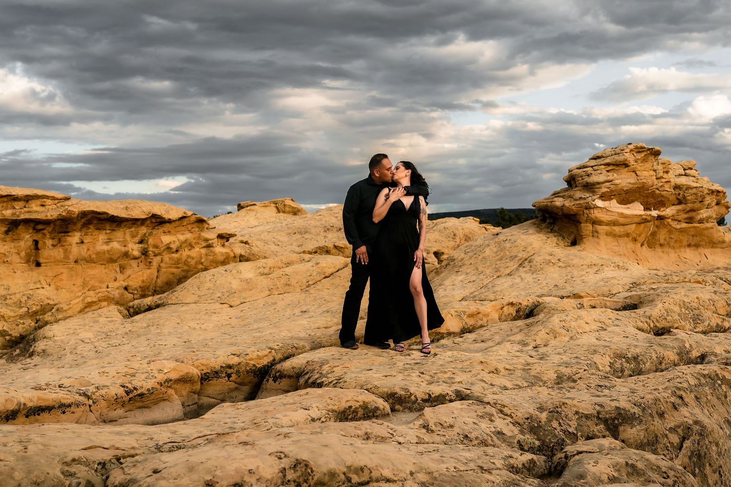 Couple dressed in black sharing a kiss among dramatic sandstone formations under a stormy sky.