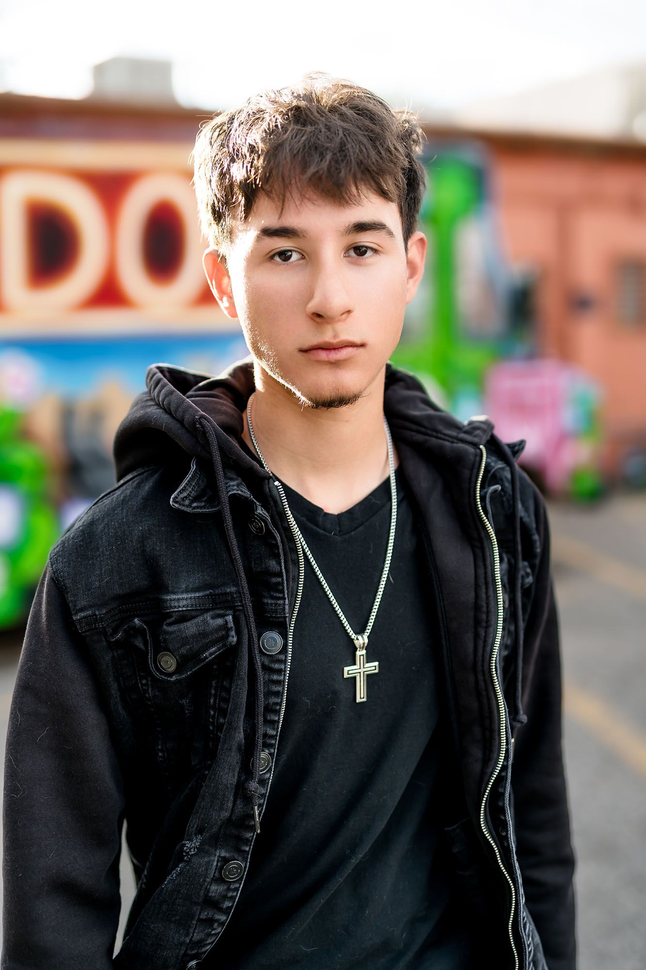 Young man in black denim jacket and cross necklace poses outdoors near colorful graffiti wall.