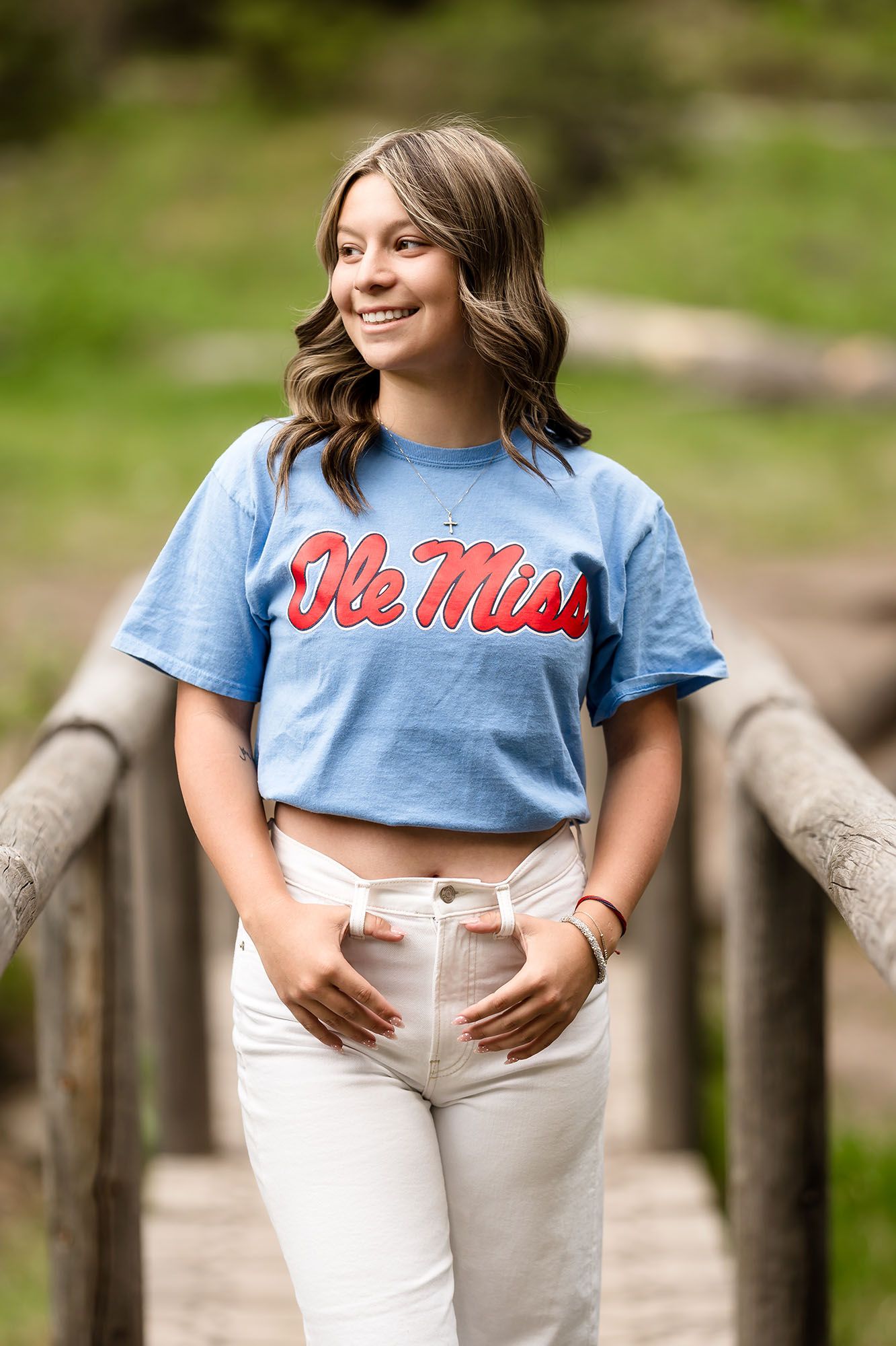 Smiling young woman wearing a light blue Ole Miss t-shirt and white jeans, standing on a wooden bridge outdoors.
