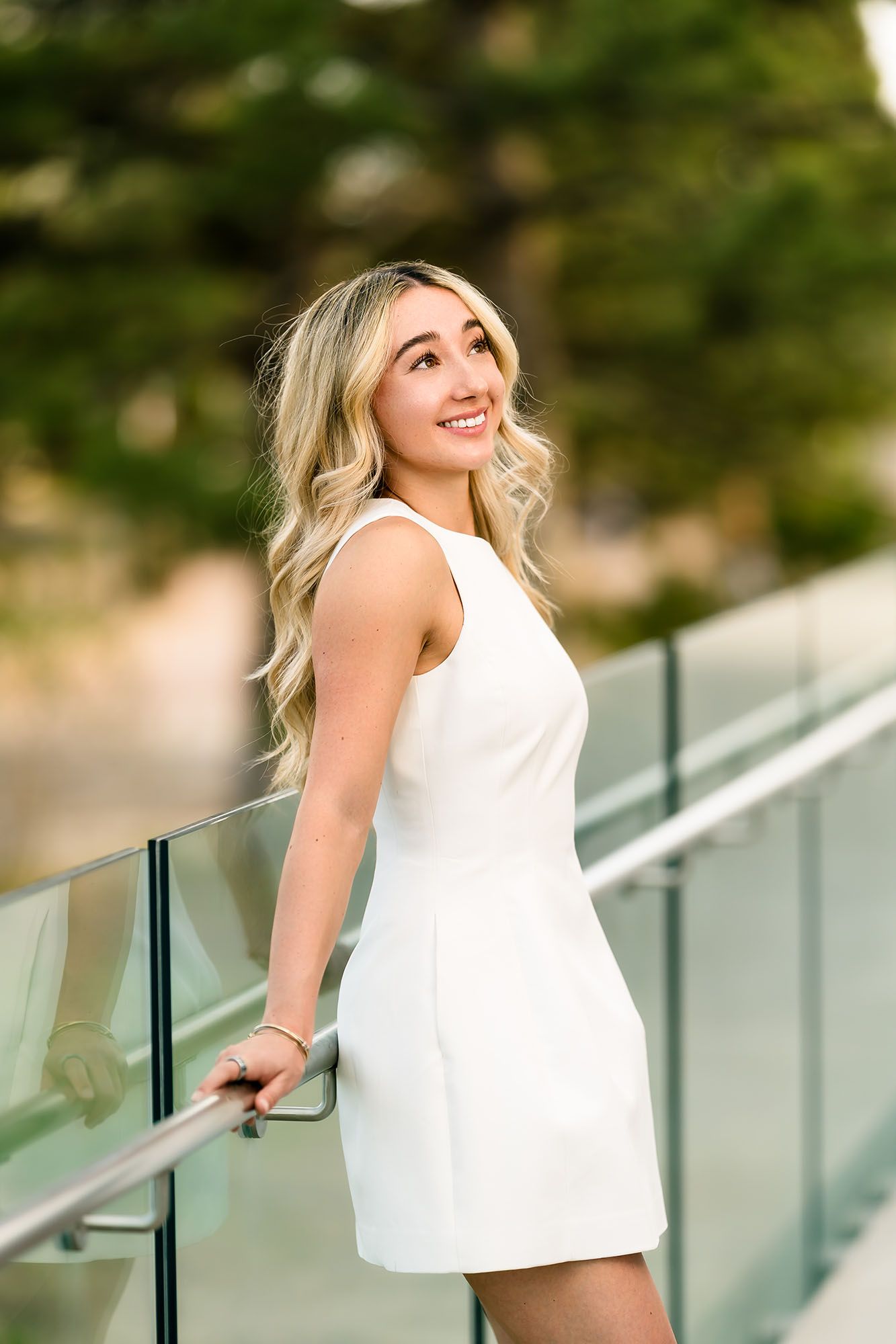 A smiling blonde woman in a white mini dress poses on a glass railing balcony outdoors.