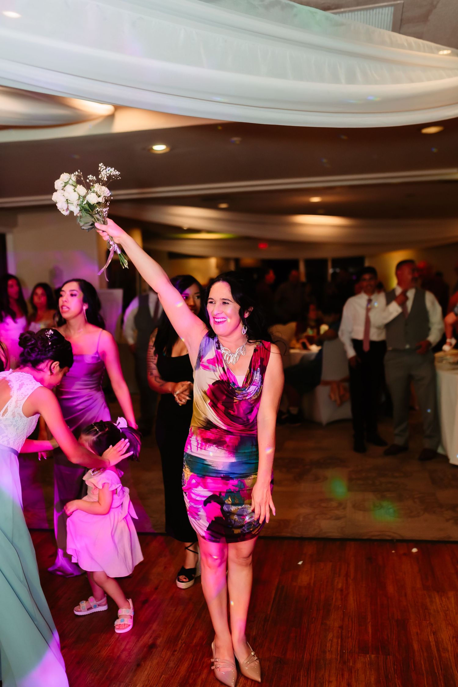Wedding guests dancing energetically under purple lighting on a wooden dance floor.