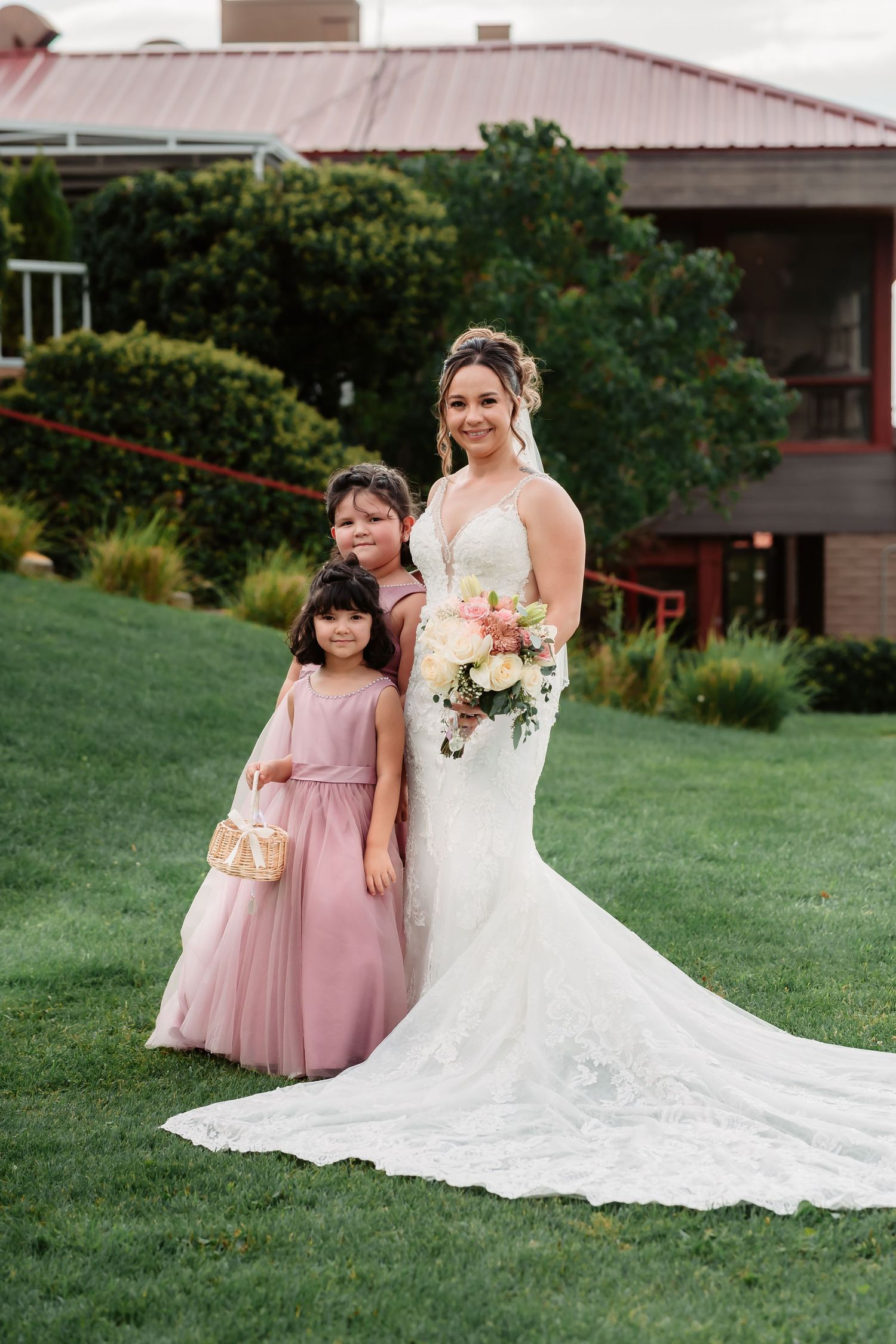 Wedding party members in formal attire pose together on green grass with a rustic building backdrop.