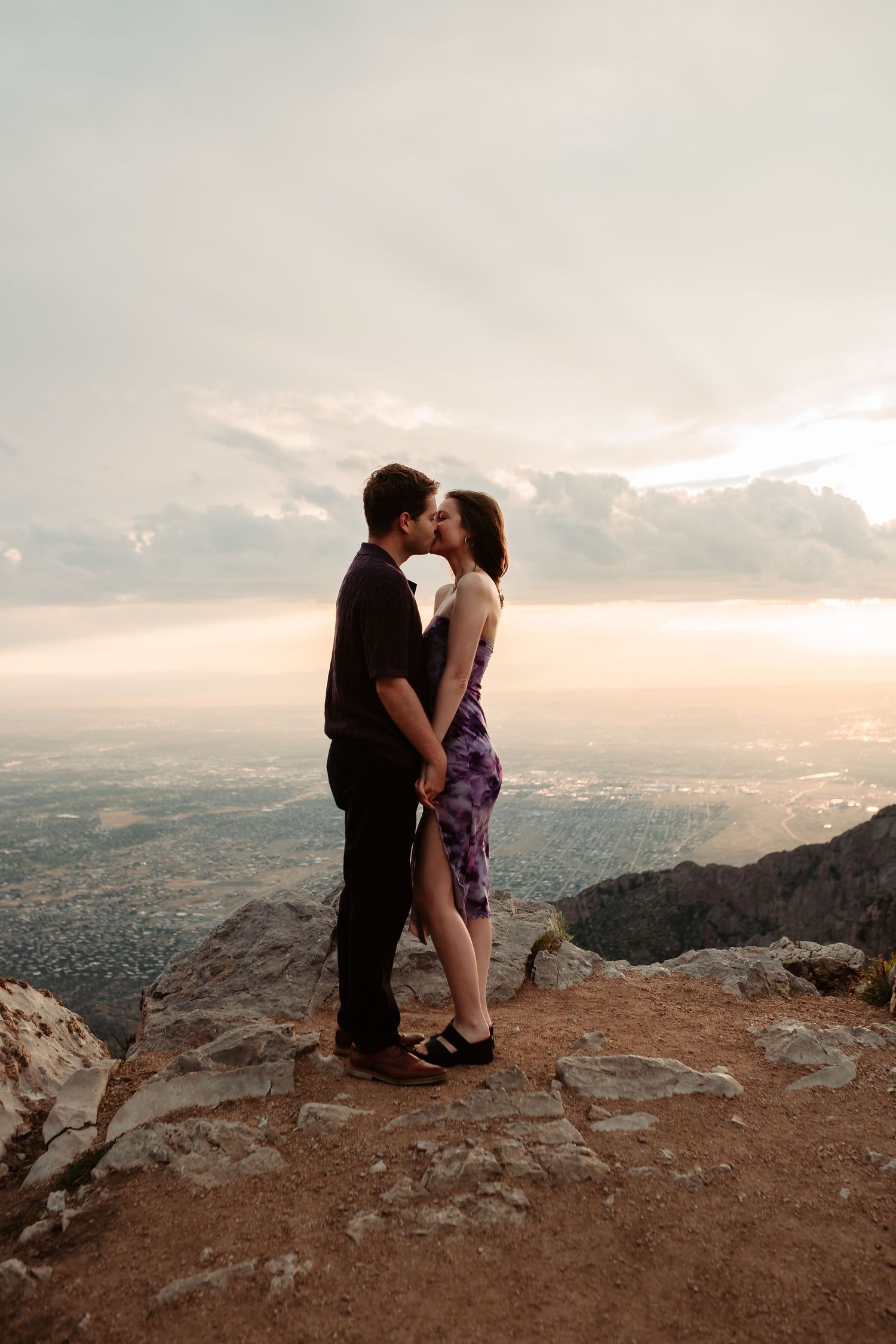A romantic sunset embrace on a mountain cliff overlooking a vast cityscape.