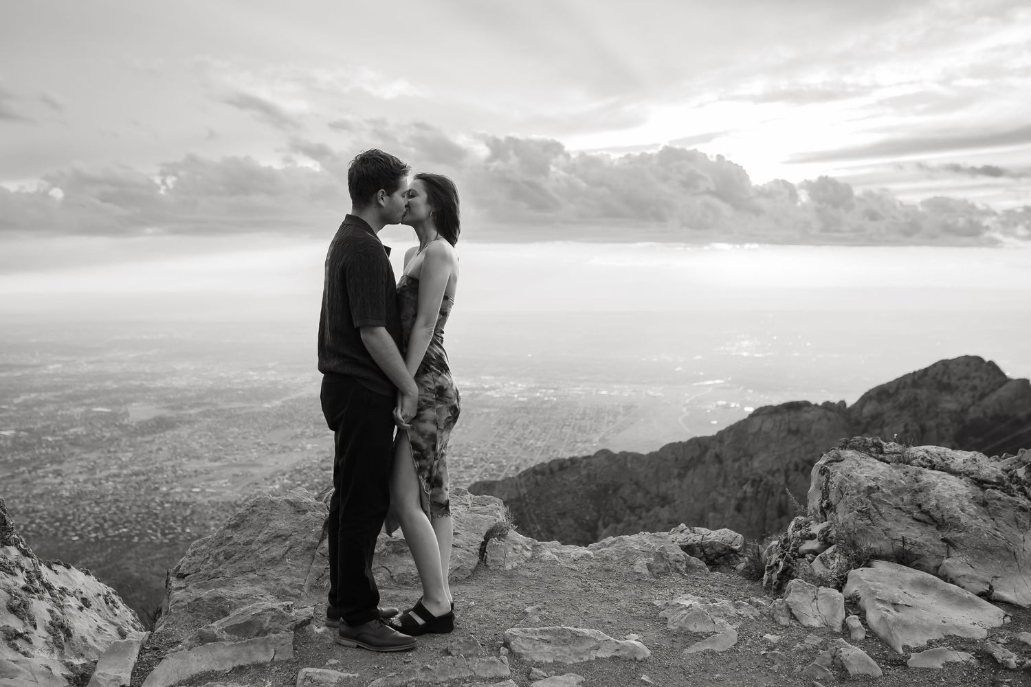A couple shares an intimate moment on a mountain overlook with a dramatic black and white landscape view.