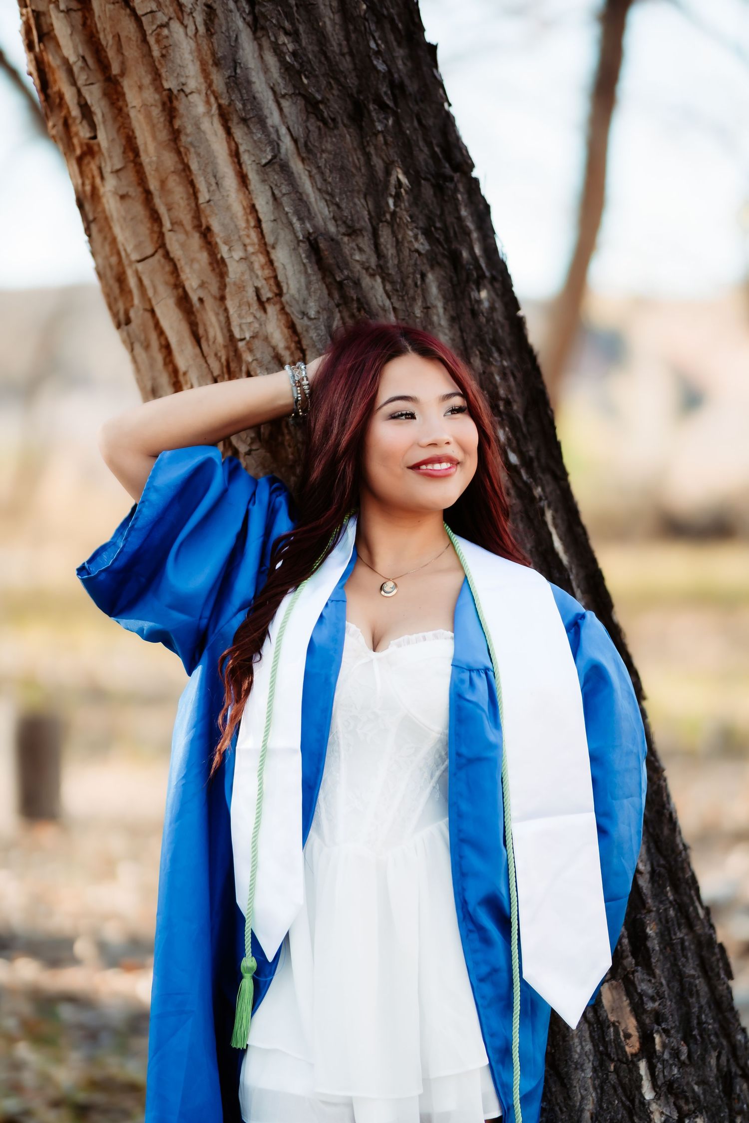 Graduate in blue cap and gown with white stole poses near tree during outdoor photo session.