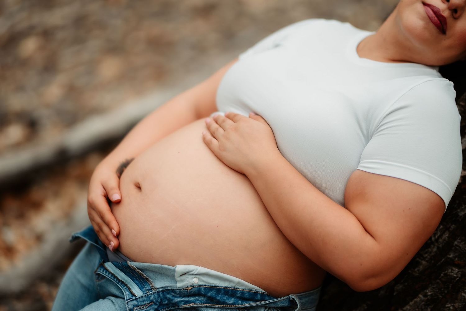 A maternity photoshoot shows a woman in a white dress resting on autumn leaves in a natural outdoor setting.