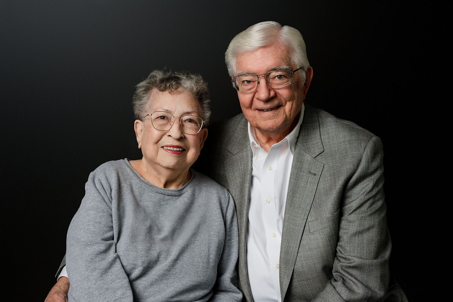 Elderly couple in casual attire pose together for a series of candid portrait photographs against dark background.
