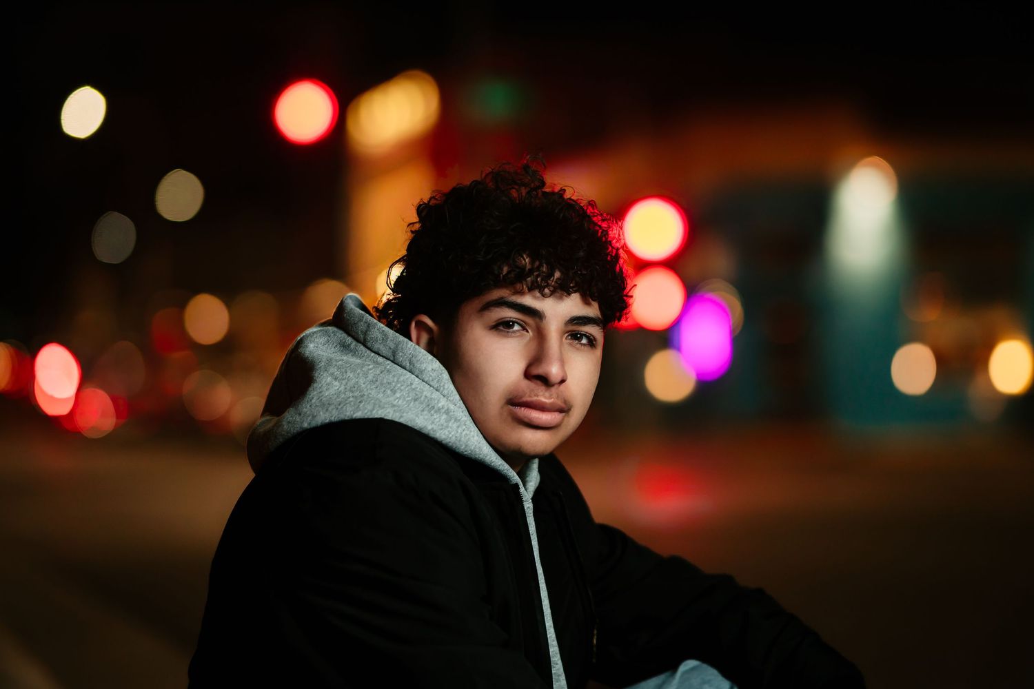 Silhouetted figure wearing a hoodie stands against a blurred background of colorful bokeh lights at night.