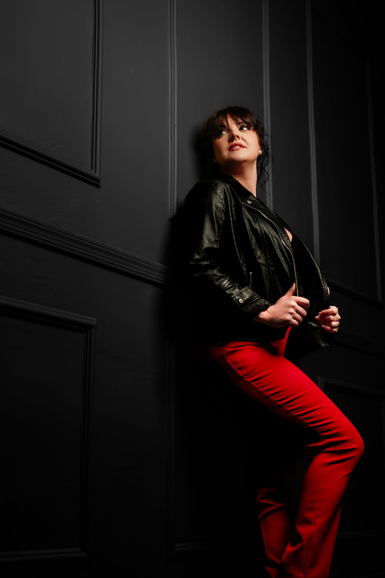 Dramatic fashion portrait showing leather jacket and bright red pants against dark moody background.