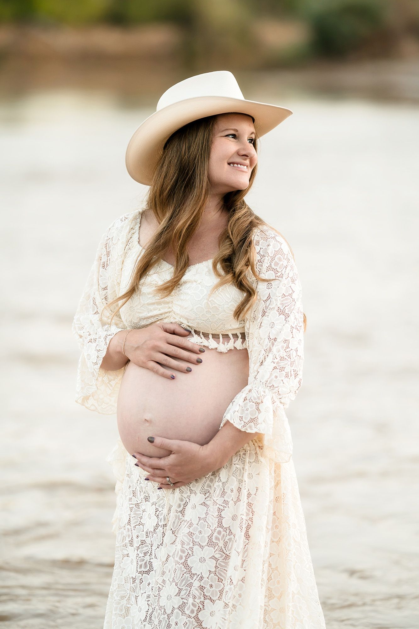 Smiling pregnant woman in white lace dress and hat cradling baby bump outdoors near water.