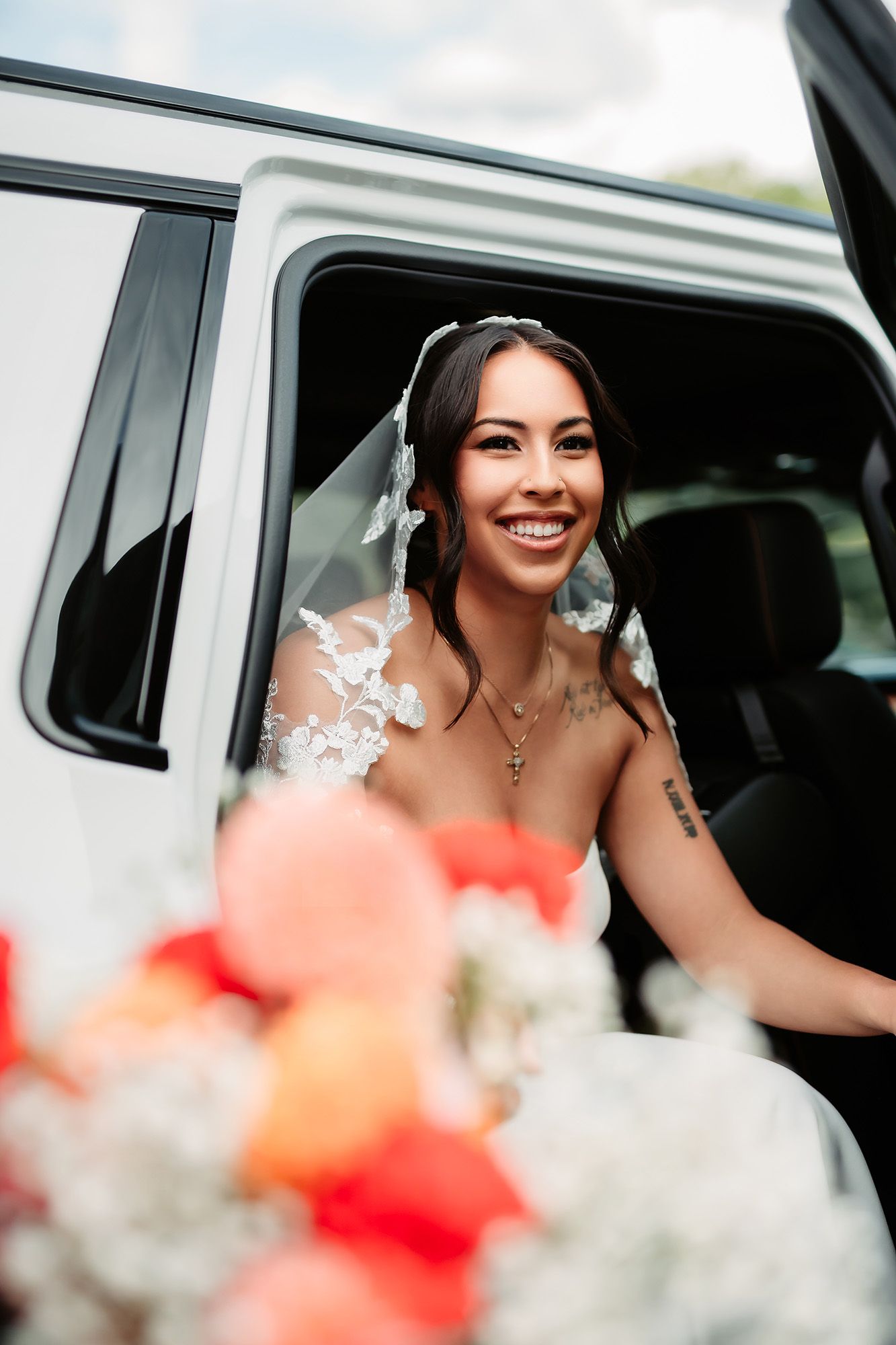 A bride smiles joyfully while sitting in a wedding car with blurred red and white flowers in the foreground.