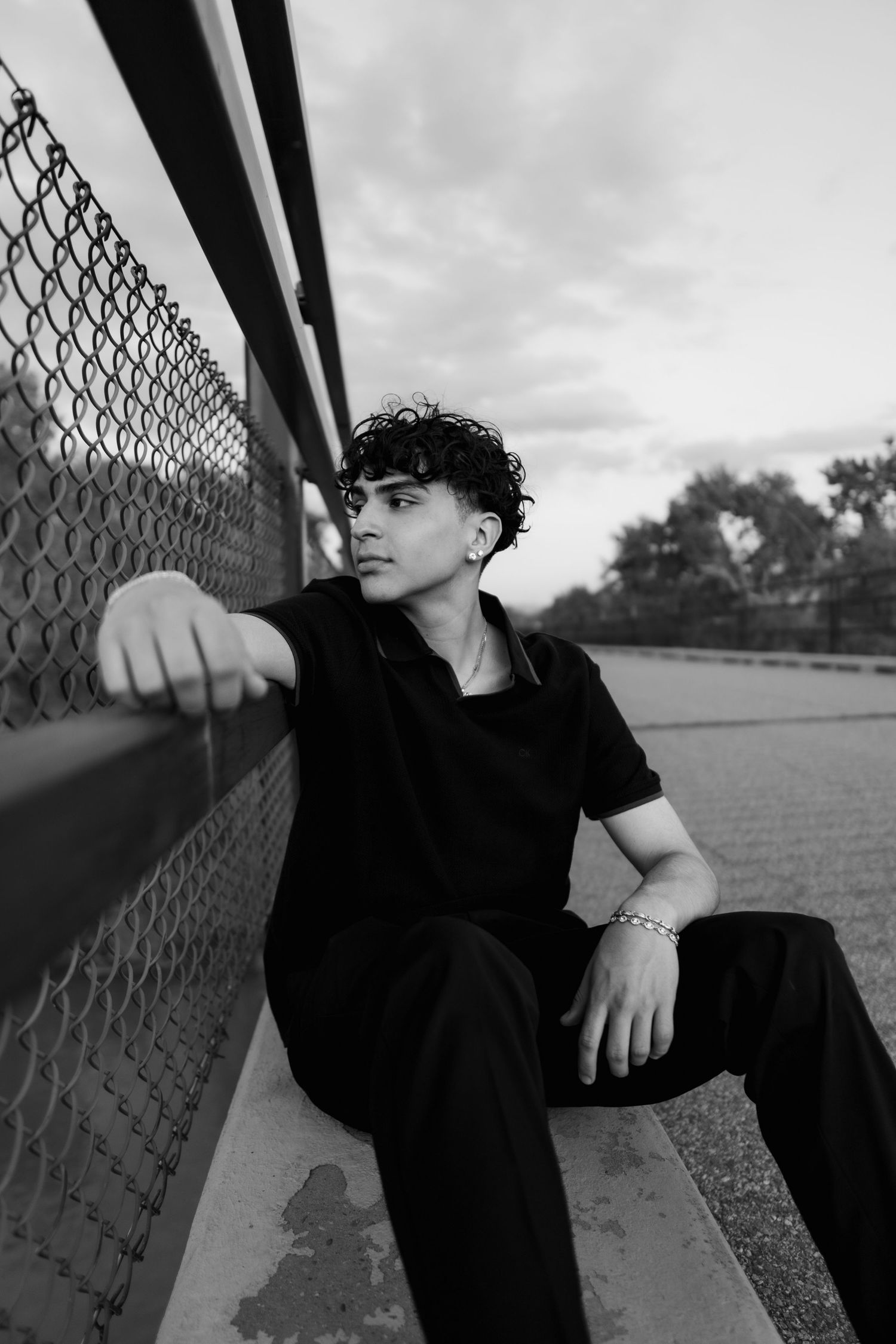 A moody black and white portrait of someone sitting against a chain link fence on an overcast day.