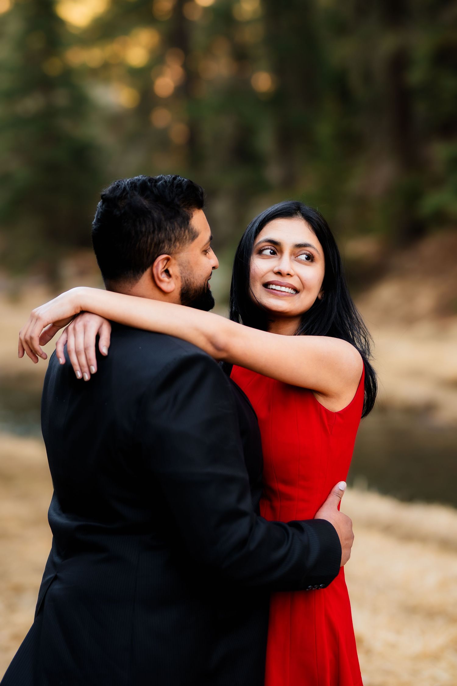 A couple embracing and smiling while the woman wears a vibrant red dress in nature.