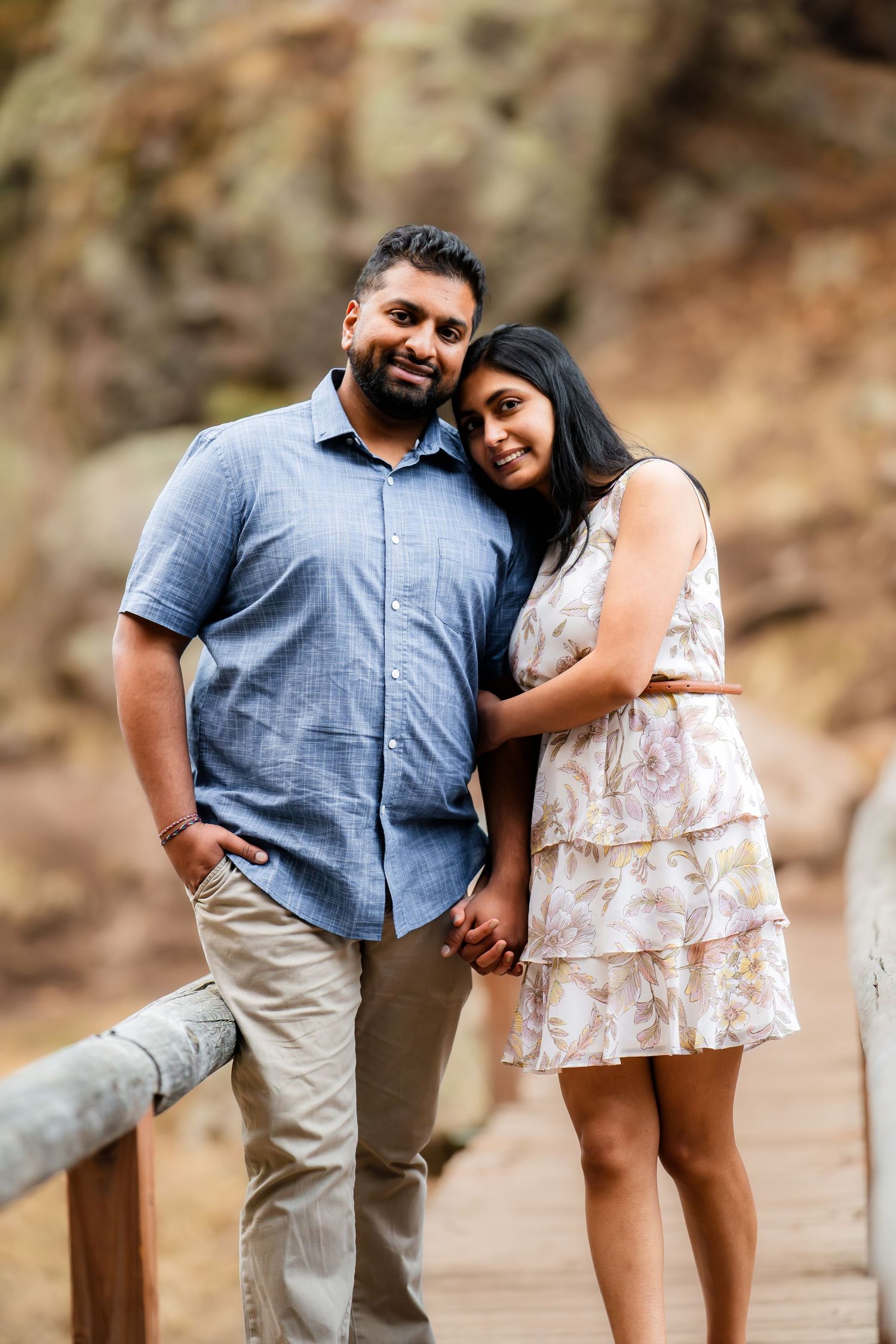 A couple in casual attire poses together on a wooden walkway with rocky cliffs in the background.