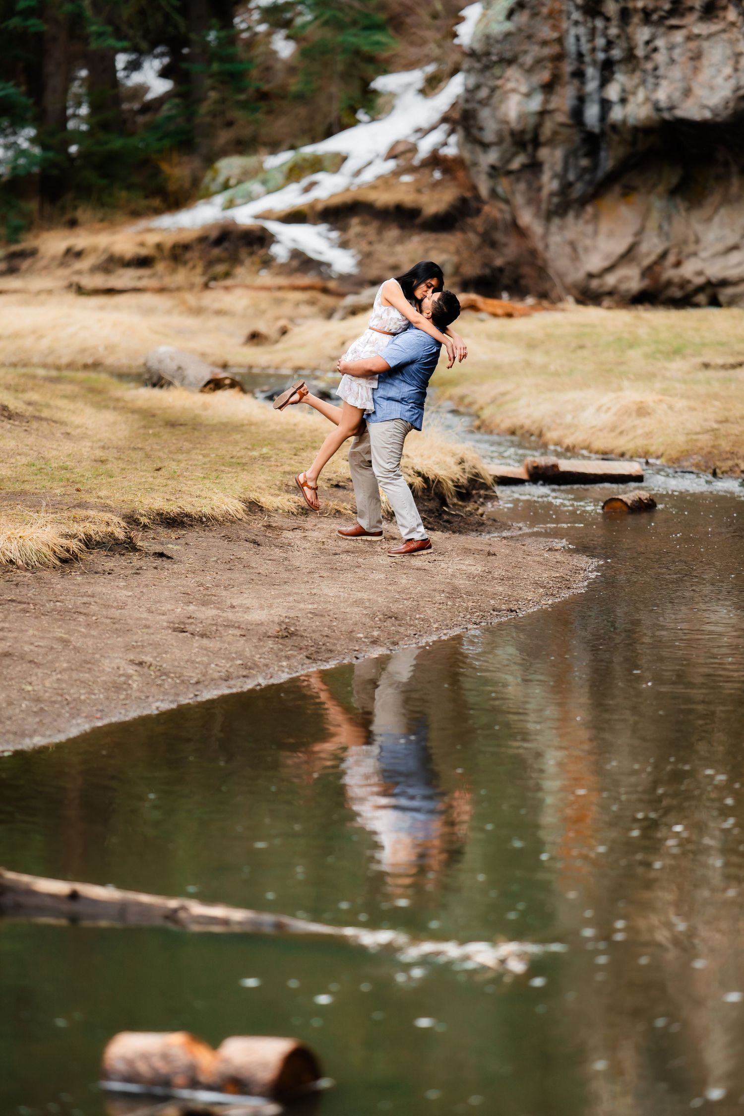 A couple shares a playful moment near a reflective pond with cliffs behind them.