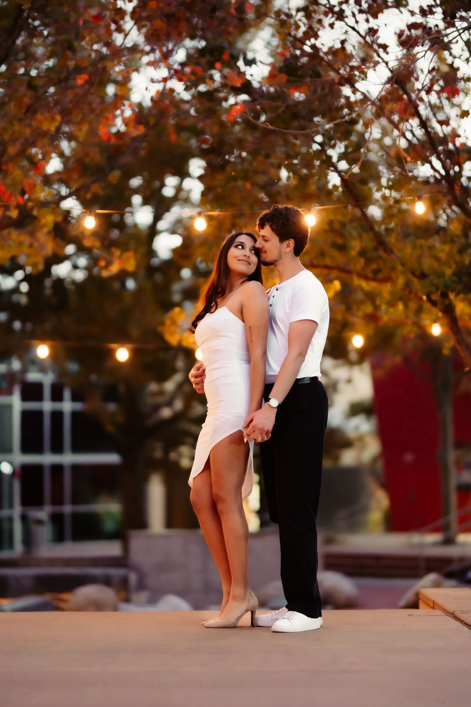 A couple poses romantically under autumn trees with string lights creating a warm ambiance.