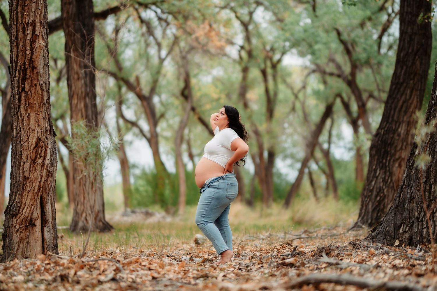 A pregnant woman stands joyfully among tall trees in a forest setting with fallen autumn leaves.