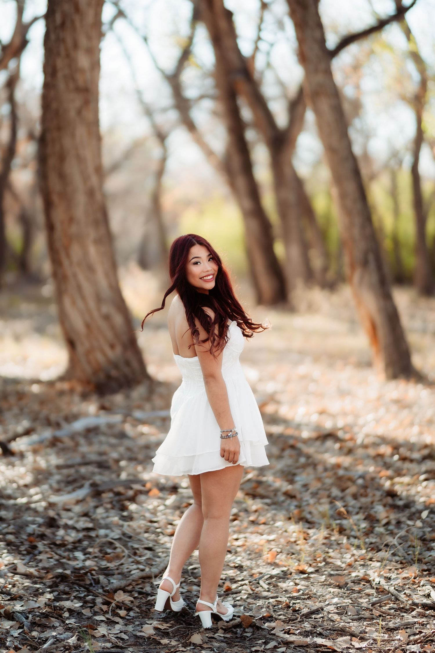 A woman in a short white dress twirls gracefully among tall trees in a forest setting.
