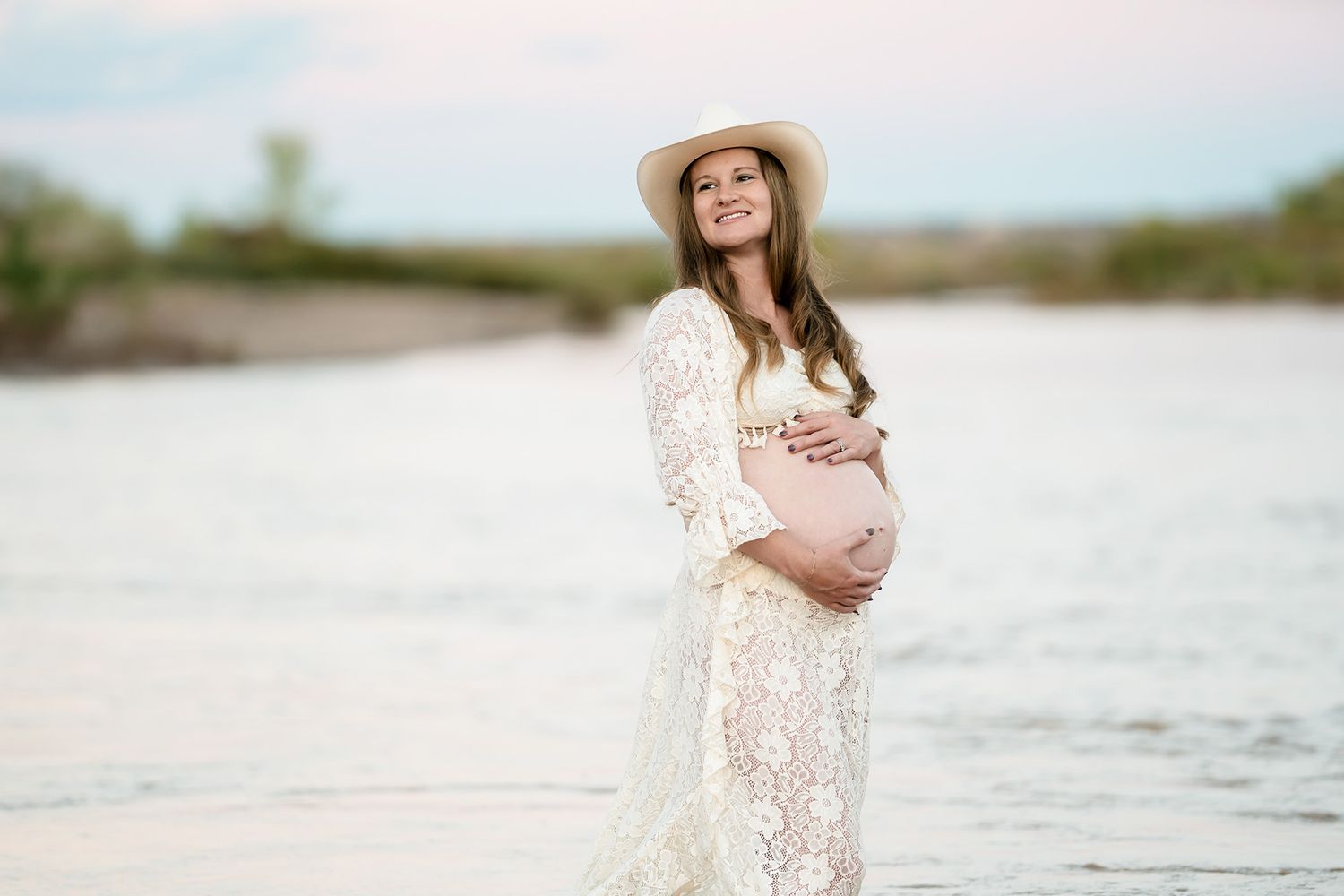 Smiling pregnant woman in white lace dress and hat cradling baby bump near a serene river.