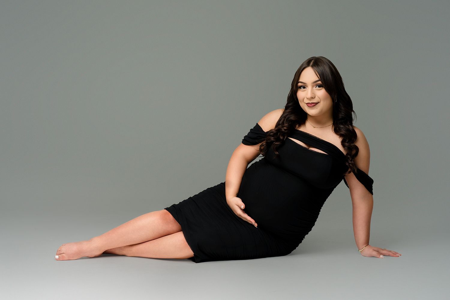 Pregnant woman in black off-shoulder dress poses gracefully on studio floor against gray background.