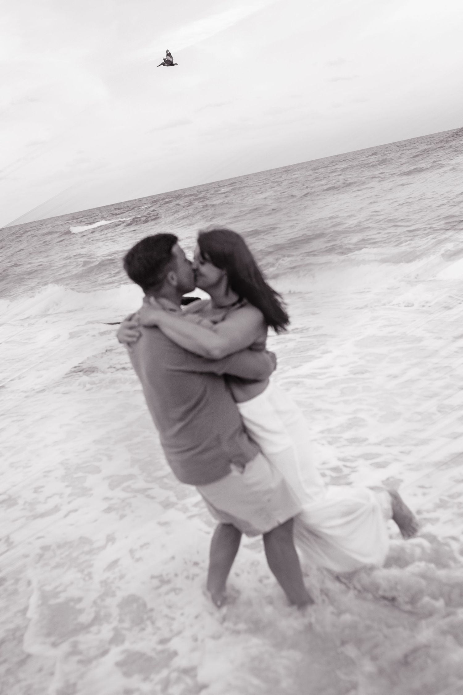 Two figures sharing an intimate embrace against ocean waves crashing on the beach in a romantic black and white scene.